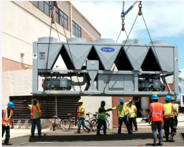 A group of several HVAC installation professionals lower a very large piece of equipment by crane into position outside an industrial building in Florida.